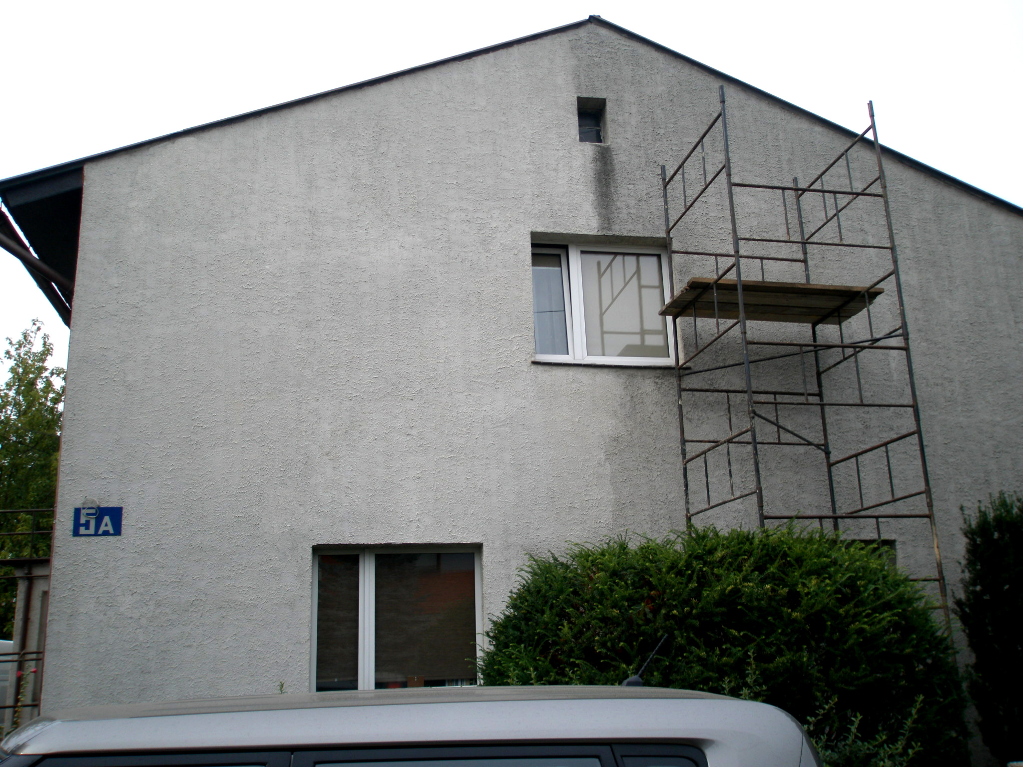 Dwupiętrowy budynek z szarą elewacją o nierównej fakturze, z widocznymi śladami zabrudzeń pod małym oknem na górze, metalowe rusztowanie przy ścianie, częściowo zasłonięte przez krzew,...